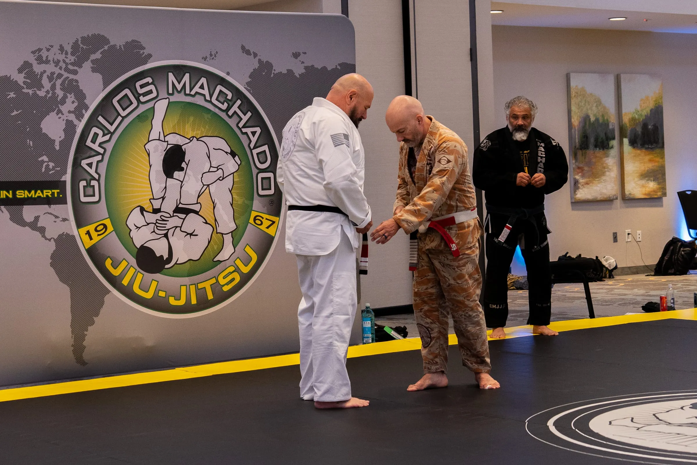 A black belt presenting a new red-tipped belt to a recipient at the Machado banner