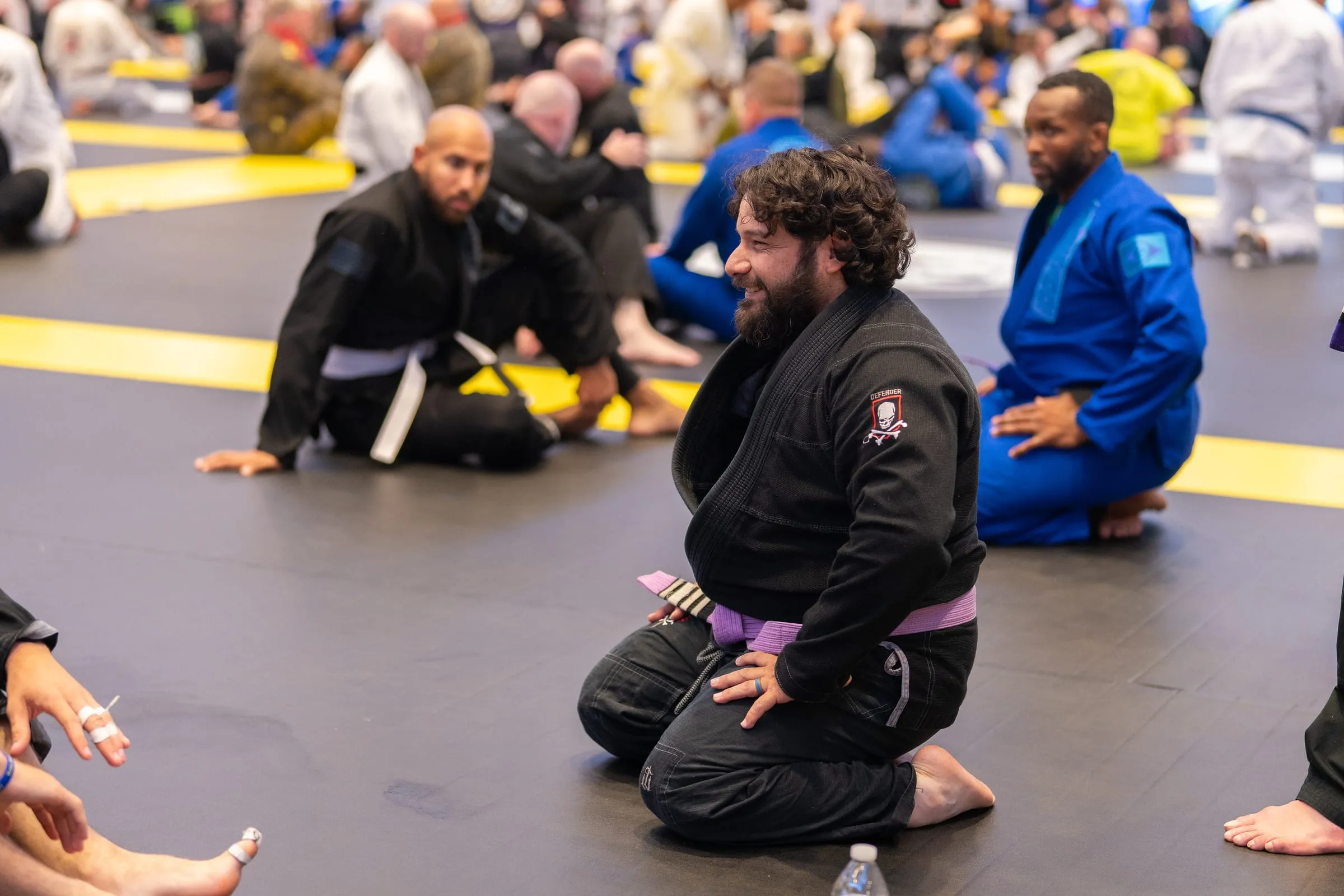 A purple belt in a black gi kneeling at the edge of a busy mat