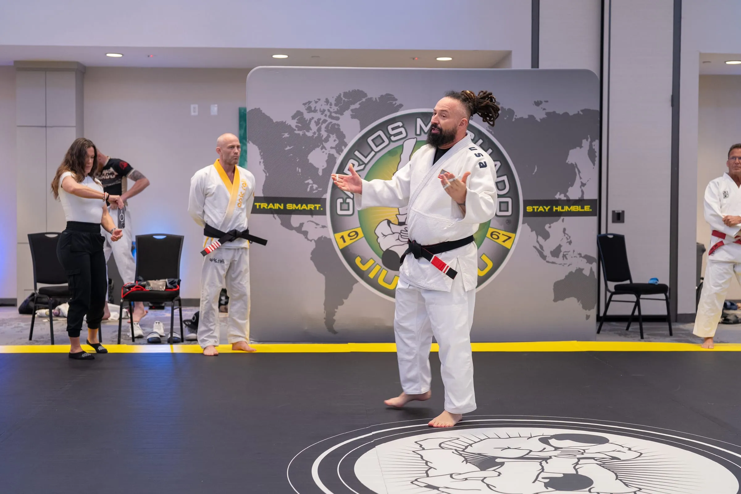 A coral-belt instructor in a white gi teaching from the centre of the mat logo