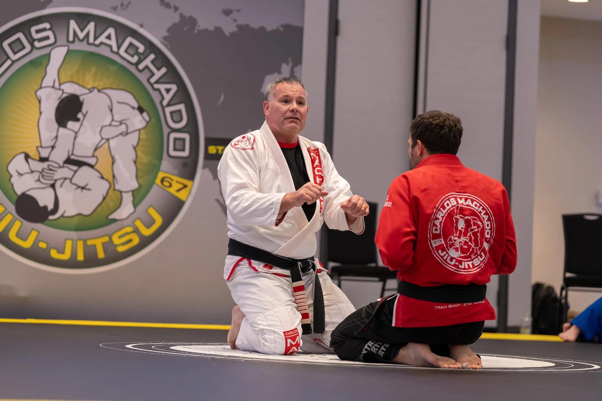 A senior black belt kneeling on the mat to teach a student in a red Carlos Machado gi