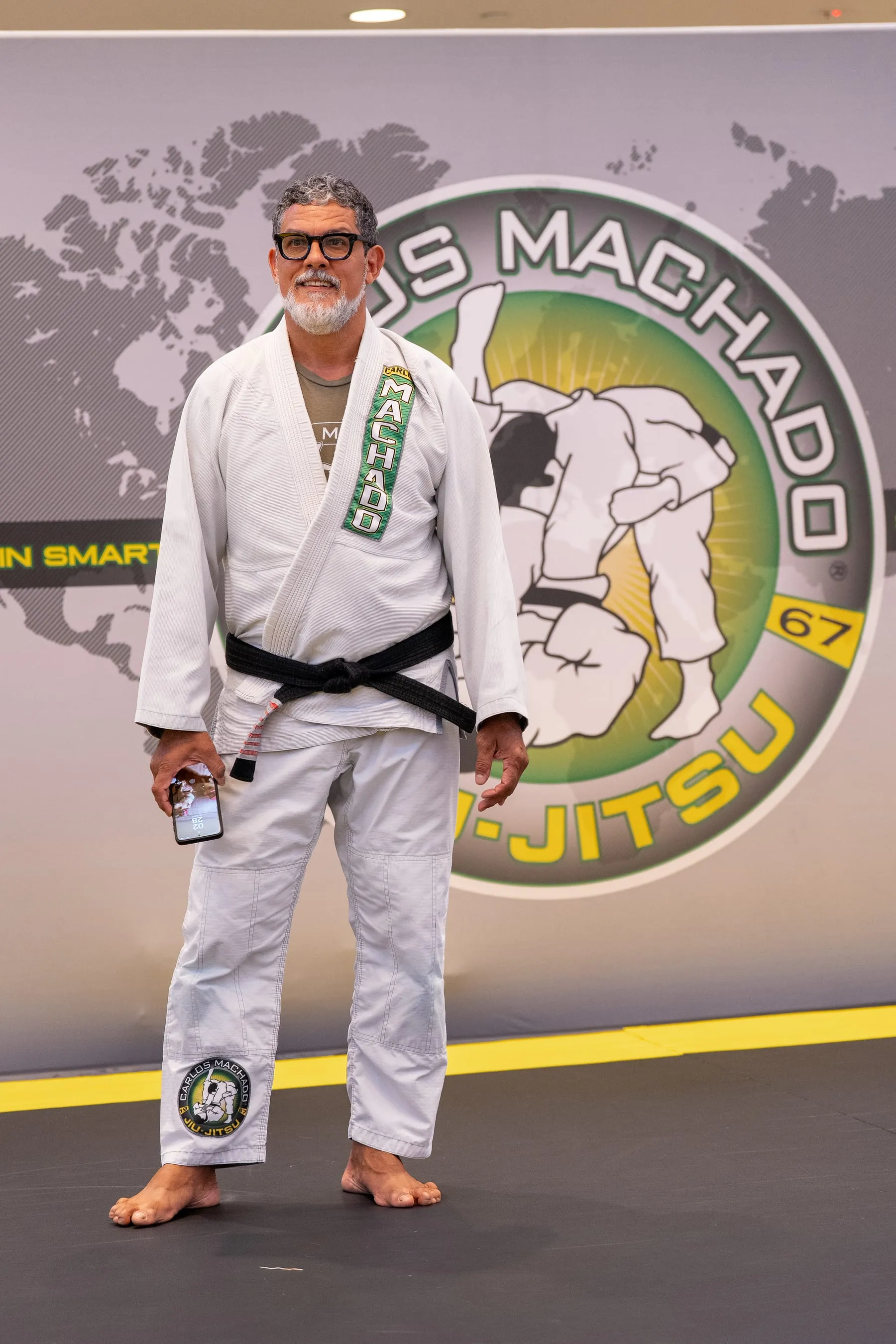 A grey-bearded black belt in a white Machado gi standing in front of the Carlos Machado banner