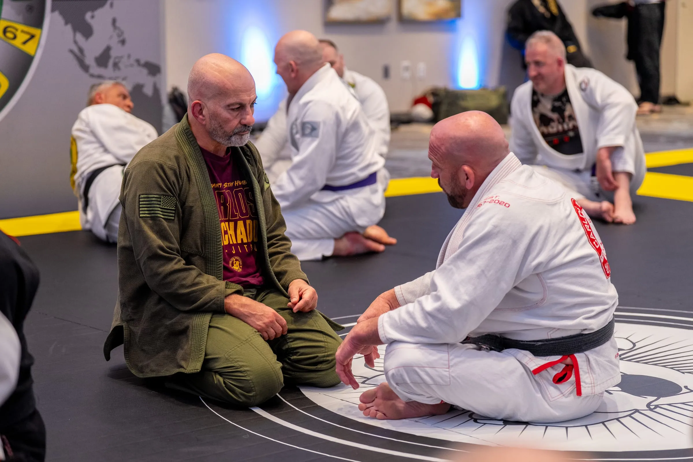 A grandmaster and a promotion recipient kneeling face to face on the mat logo